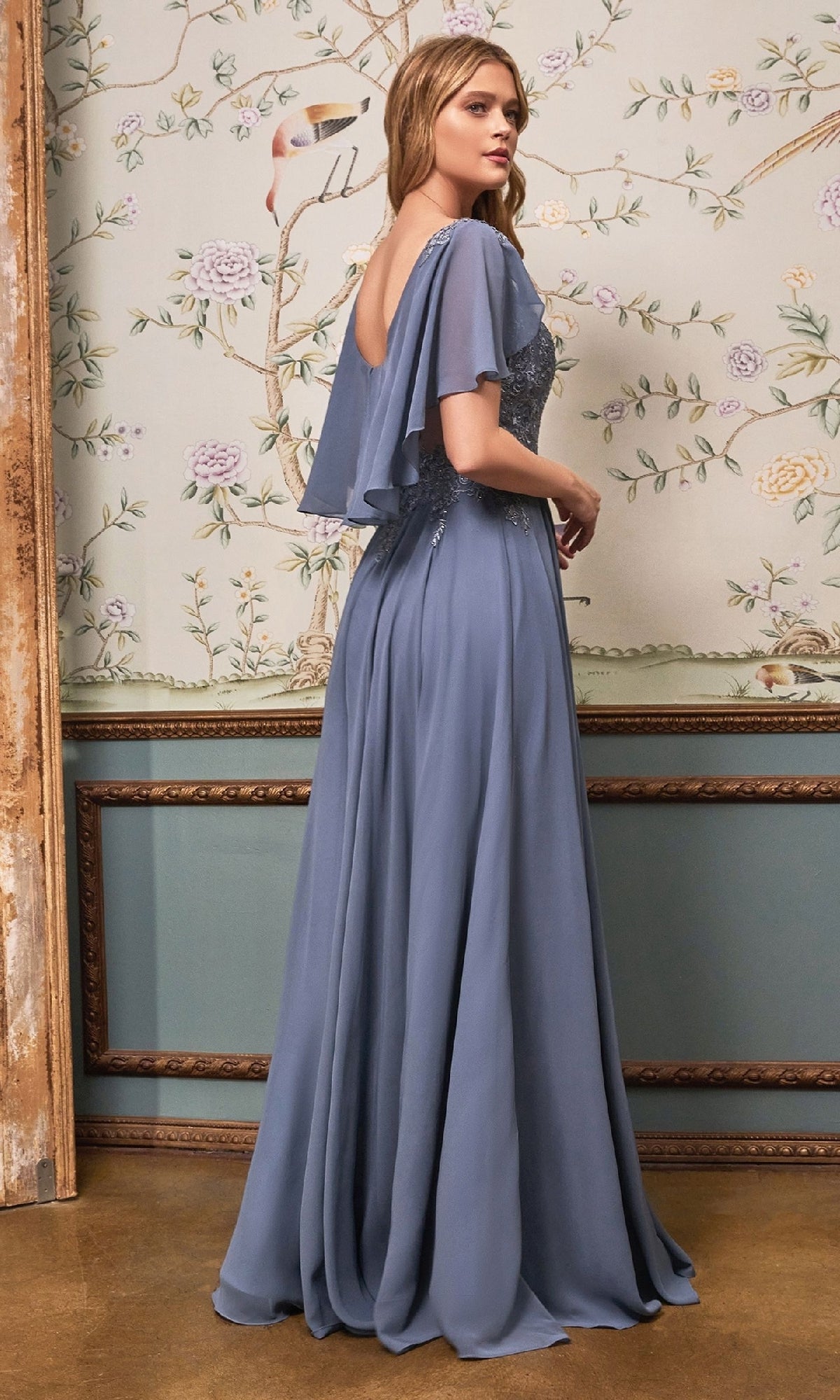 A woman wearing the Ladivine HT101 Cape-Sleeve Long A-Line Prom Dress in dusty blue chiffon stands before a floral-patterned wall, turned slightly away from the camera.